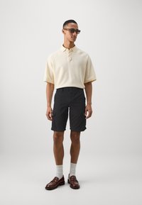Vaaleanbeige lyhythihainen polo paita, jossa on teksturoitu kuvio, yhdistettynä mustiin cargo-shortsit, valkoisiin sukkiin ja ruskeisiin loafer-kengiin.