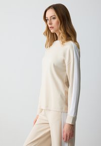 Felpa beige con accenti bianchi sulle spalle, maniche lunghe e colletto a giro. Texture morbida e vestibilità rilassata, abbinata a pantaloni beige coordinati.