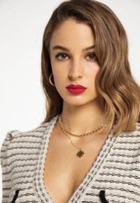 Striped cardigan dzianinowy z dekoltem w serek, wykonany z teksturowanej tkaniny. Złote łańcuszki biżuterii nałożone na szyję; odważna czerwona szminka podkreśla usta.