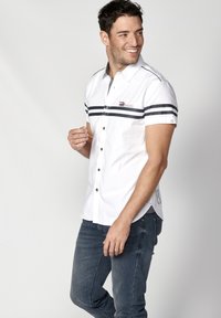 Camisa blanca de manga corta con rayas horizontales negras, un frente con botones y un acento de logo en el lado izquierdo del pecho. Combinada con jeans oscuros.