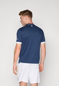 Polo deportivo en azul marino con mangas cortas acentuadas en blanco y un logo en el brazo. Presenta un cuello clásico y un diseño elegante y ajustado.
