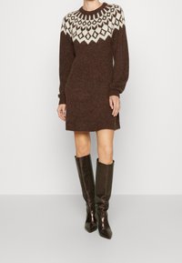 Robe en tricot marron avec un motif géométrique blanc au niveau de l'empiècement, dotée de manches longues et d'une coupe droite, associée à des bottes brillantes marron arrivant au genou.