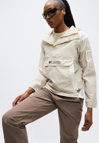 Γυναίκα που φοράει μπεζ μπουφάν Columbia Sportswear, καφέ παντελόνι και σκουρόχρωμα γυαλιά ηλίου, μπροστά σε λευκό φόντο.