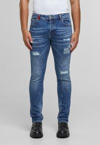 Jeans de mezclilla azul con corte ajustado, diseño desgastado, parches desgastados y detalles de salpicaduras de pintura. Presentan un acento rojo en la cinturilla.