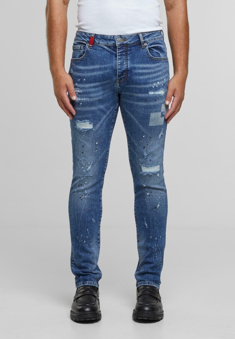 Jeans de mezclilla azul con corte ajustado, diseño desgastado, parches desgastados y detalles de salpicaduras de pintura. Presentan un acento rojo en la cinturilla.