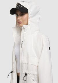 Jachetă albă de ploaie cu glugă, dotată cu șnururi ajustabile, accente negre și detalii de design perforat pe față.