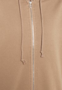 Gros plan d'un sweat à capuche beige zippé montrant une fermeture éclair argentée et deux cordons aux extrémités effilochées.