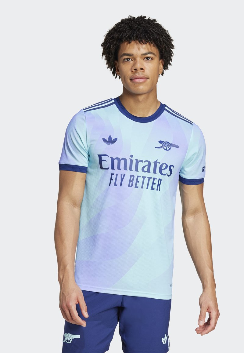 Lys blå fodboldtrøje med mørkeblå accenter, der har logoet "Emirates Fly Better" og tre striber på skuldrene.