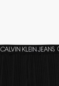 Czarny pas z białym napisem "CALVIN KLEIN JEANS" na plisowanej czarnej tkaninie poniżej.