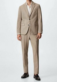 Completo beige con una vestibilità su misura, giacca a un bottone, rever classico e due tasche frontali. Abbinato a una camicia bianca e mocassini marroni.