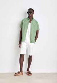 Cămașă verde cu mânecă scurtă și nasturi, cu un buzunar, top alb fără mâneci, pantaloni scurți albi și sandale slip-on bej cu accente negre.
