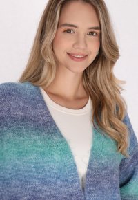 Cardigan lavorato a maglia in sfumature di blu e verde, con scollo a V, texture morbida e design aperto. Indossato sopra una maglietta bianca.