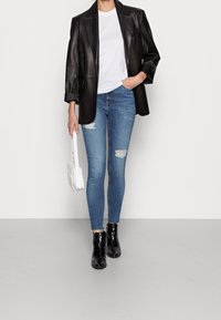 Blazer de cuero negro sobre una camiseta blanca, jeans ajustados azules desgastados, botas negras hasta el tobillo y un pequeño bolso blanco.