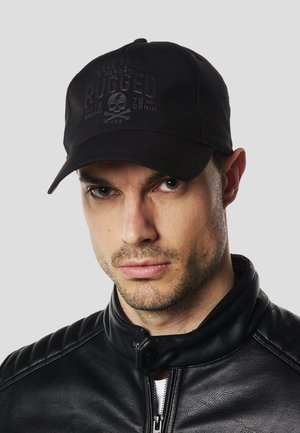 Hombre con una gorra negra de béisbol con un logo de calavera y el texto "RUGGED", combinado con una chaqueta de cuero negra, fondo gris neutro.