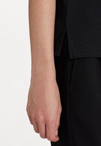 Svart ribbad t-shirt med sidoflikar och broderad logodetalj. Hand synlig, vilande bredvid ett svart plagg.