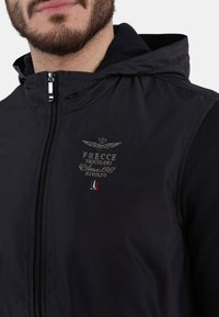 Gilet nero con cappuccio, dotato di zip, logo ricamato con aquila e dettagli della bandiera italiana. Esterno liscio con maniche texture.