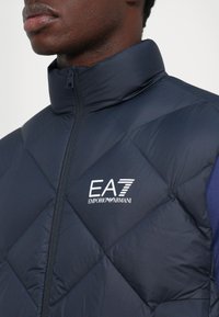 Gros plan sur une personne portant une veste matelassée bleu marine avec un col haut et un logo EA7 Emporio Armani blanc sur la poitrine.