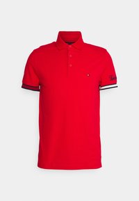 Rød polo skjorte med kraveformet halsudskæring, tre knapper og korte ærmer. Har marineblå og hvide stribede detaljer på ærmerne og broderet logo.
