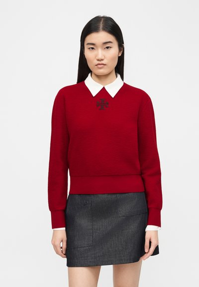 Woman with long black hair wearing a red sweater with white collar and a dark gray skirt, standing against a plain light background.