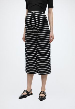 Femme portant un pantalon large court rayé noir et blanc avec un top noir à manches courtes et des ballerines noires pointues à brides.