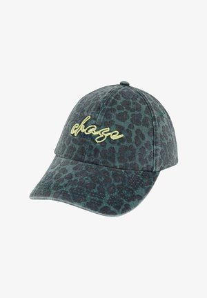 Kvinde iført en gul kortærmet skjorte, blå jeans og en mørkegrøn kasket med gul skrift, stående med den ene hånd i lommen.