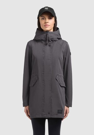 Nő hosszú, sötétszürke kapucnis dzsekiben, fedeles zsebekkel és fekete baseballsapkában, világos egyszínű háttér előtt állva.