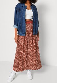 Jeansjacke mit hochgerollten Ärmeln über einem weißen Cropped-Top, kombiniert mit einem braunen, blumigen Maxirock und weißen Sneakers. Strukturierte Muster sind sichtbar.