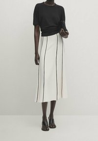 Schwarzes T-Shirt kombiniert mit einem weißen Midirock mit vertikalen schwarzen Streifen. Dazu schwarze Sandalen und eine kleine schwarze Tasche in der Hand.