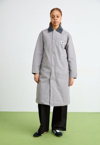 Cappotto lungo grigio con colletto morbido, tasche frontali e chiusura a bottoni. Abbinato a pantaloni neri a gamba larga e pantofole scure.
