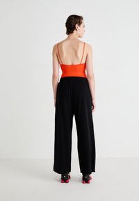 Red fitted bodysuit with thin straps paired with loose black pants. Black shoes with pink accents. Clean, minimal background setting.