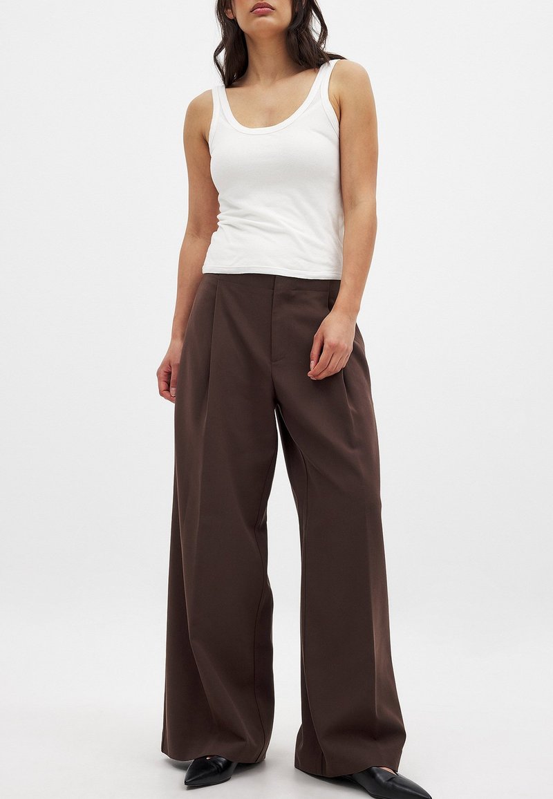 Pantalones de pierna ancha marrones con una textura suave, que presentan una cintura alta y un pliegue planchado; combinados con una camiseta sin mangas blanca. Zapatos de punta negra.