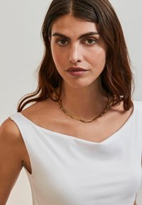 Vit ärmlös topp med en mjukt draperad halsringning, prydd med ett guldhalsband med avlånga länkar. Slät textur och minimalistisk design.