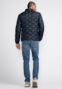 Marineblauer, gesteppten Pufferjacke mit hohem Kragen, die eine glänzende Textur und elastische Bündchen aufweist. Getragen mit hellblauen Jeans und lässigen Sneakers.