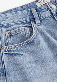 Jeans in denim azzurro chiaro con rivetti metallici, tasca anteriore, passante per cintura, bottone ed etichetta con la scritta "Wide Leg High Rise, Made in Bangladesh."