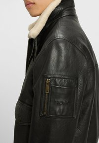 Czarna skórzana kurtka z owczym kołnierzem, z zamykaną kieszenią na lewym rękawie i wytłoczonym logo. Widoczna fakturowana powierzchnia.