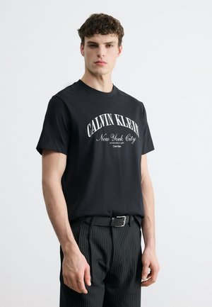 Νέος άνδρας που φοράει μαύρο μπλουζάκι Calvin Klein με την επιγραφή "New York City" και μαύρο παντελόνι με λεπτές ρίγες, σε απλό ανοιχτό φόντο.