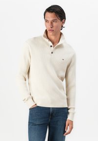 Homme aux cheveux foncés portant un pull crème à côtes avec des boutons et un jean bleu foncé, se tenant avec une main dans la poche.