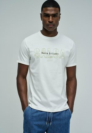 Hombre con camiseta blanca con el texto "SALSA Denim Artisans Jeans" y pantalones vaqueros azules, de pie con las manos en los bolsillos.