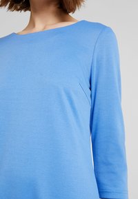 Blauwe lange mouwtop met een wijde bootnek. Zachte, gladde stof met een subtiele textuur. Aansluitende lijfje en iets ontspannen mouwen.