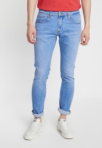 Hombre vestido con jeans slim-fit de color azul claro, remangados en los tobillos, zapatillas blancas y una camisa roja, de pie contra un fondo liso.