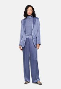 Woman with shoulder-length black hair wearing a shiny lavender suit with wide-leg pants, matching turtleneck, and gray pointed flats, standing.