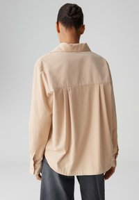 Light beige button-up shirt with a collar, relaxed fit, back pleat detail, and long sleeves. Paired with dark denim pants.