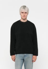 Svart sweatshirt med rund hals och texturerad frampanel, långa ärmar och löst sittande passform, tillsammans med ljusblåjeans.
