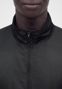 Gros plan d'une personne portant une veste noire zippée avec un col montant, montrant le bas du visage et la zone du cou.