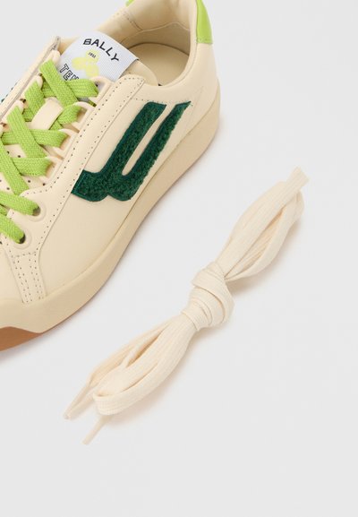 Αθλητικό παπούτσι εκρού με πράσινα κορδόνια, σκούρο πράσινο υφασμάτινο λογότυπο και ένα επιπλέον ζευγάρι δεμένα λευκά κορδόνια σε γκρι φόντο.