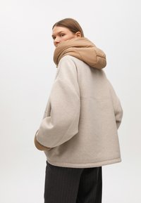 Bež jakna od flisa u oversized stilu s pufastim kapuljačom u boji sipine. Dizajn sadrži ravnu liniju kroja, spuštene ramena i dva bočna džepa.