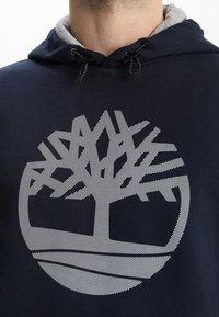 Sudadera de color azul marino con forro interior gris, que presenta un gran gráfico blanco de un árbol en el centro y detalles a rayas en todo su diseño.
