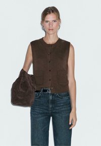 Gilet sans manches marron avec fermeture à bouton et deux poches avant, associé à un jean en denim bleu foncé et un sac en daim marron foncé.