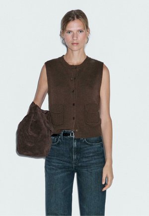 Brown sleeveless vest with button closure and two front pockets, paired with dark blue denim jeans and a dark brown suede bag.