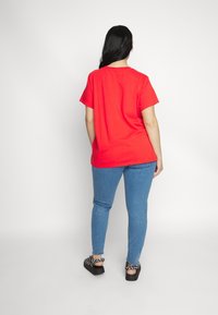T-shirt en coton rouge à manches courtes, associé à un jean skinny bleu clair et des sandales noires avec des accents décoratifs.